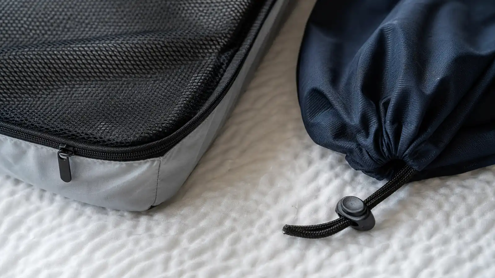 Close-up of a packing cube corner seam and a slightly loosened drawstring toggle on a laundry bag, shown side by side on a bed