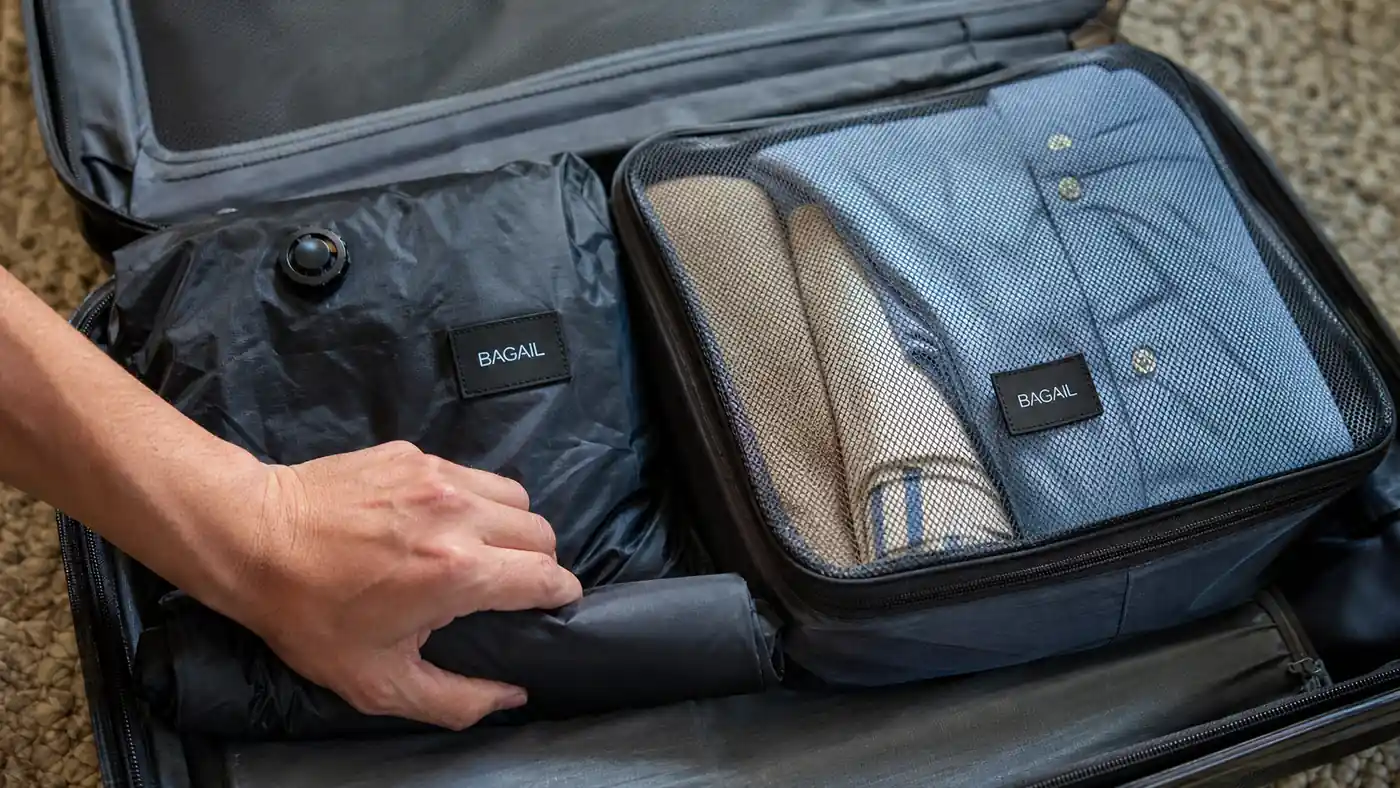 Hand pressing air out of a rolled compression bag next to an open BAGAIL packing cube filled with folded shirts