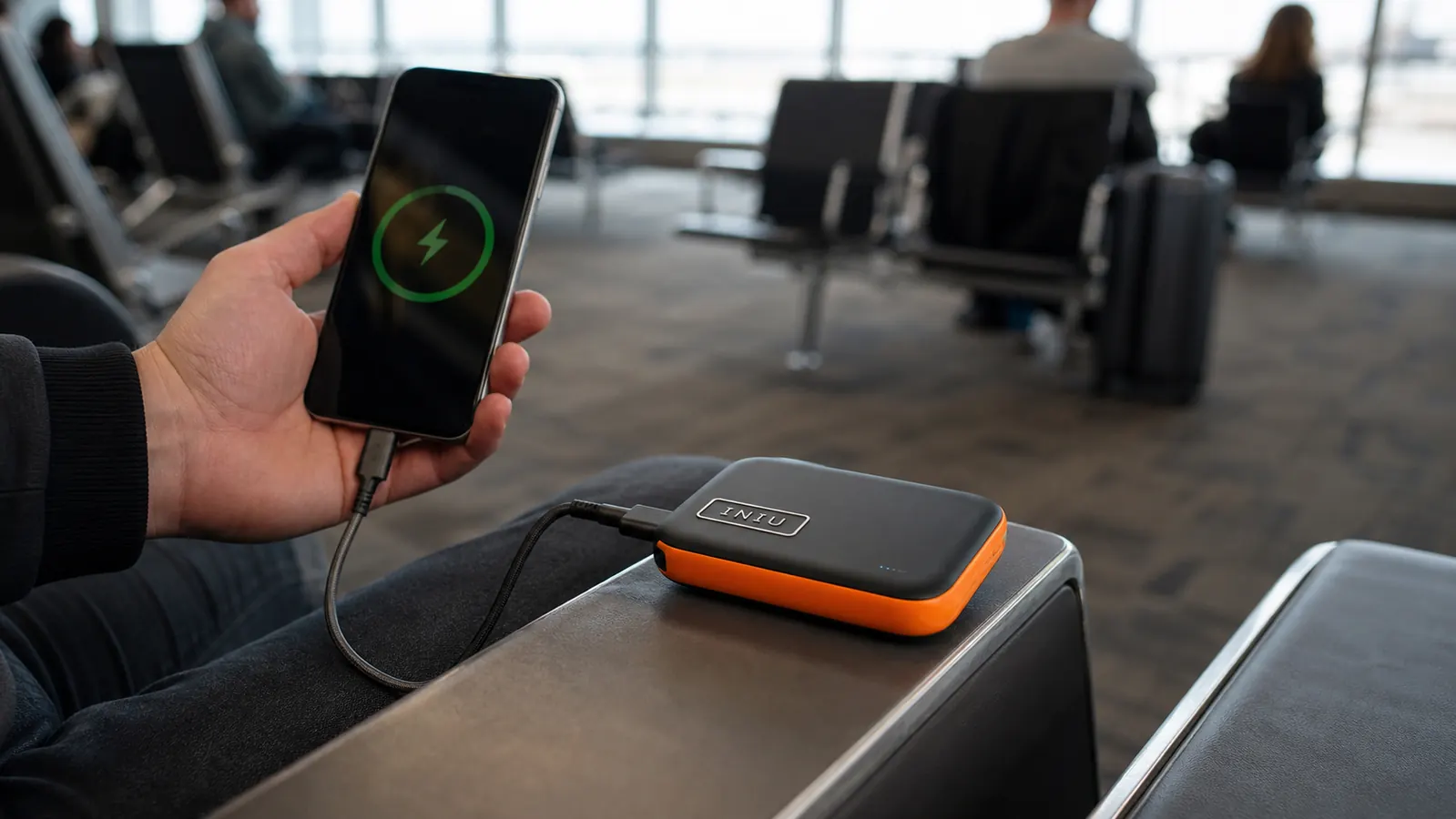 Hand holding a compact black INIU power bank plugged into a smartphone at an airport gate seat