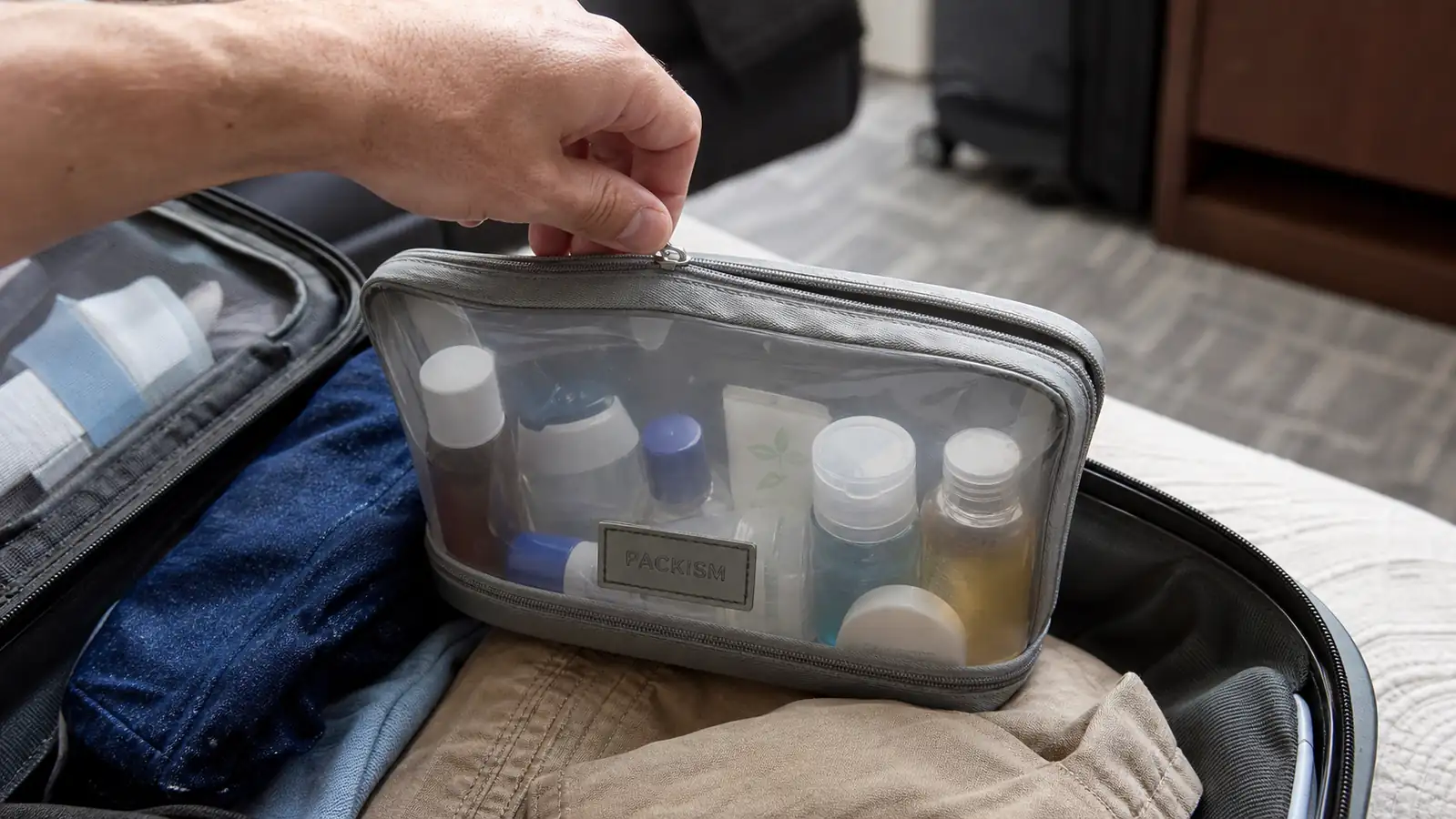 Hand zipping a clear PACKISM toiletry bag filled with travel-size toiletry bottles inside an open carry-on suitcase
